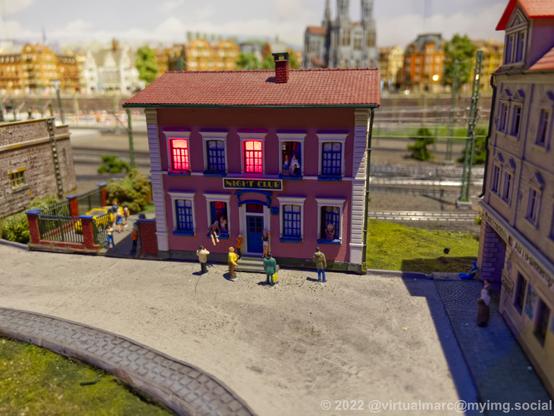 A photo of a building from a model railway, labled with "Night club". Red light is shining from the inside, it's a brothel. Some womone staning in the windows, some men standing in front of it.