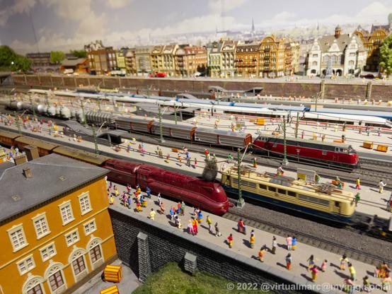 A photo of a big train station from a model railway, with passenger trains from different times, including a steam locomotive and an ICE. Two fright trains, a diesel and an electric, a driving through the station. There are hundreads of people on the platforms. A city can be seen in the background.
