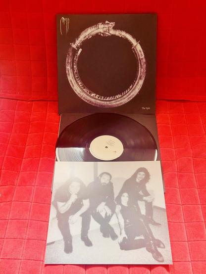 Vinyl cover of 'The Spin' by Messa. The artwork features a dark background with a glowing circular snake eating its own tail (ouroboros), and the band's name in a wispy font in the top left. The vinyl is partially out, and a black-and-white photo insert shows the four band members seated, looking at the camera