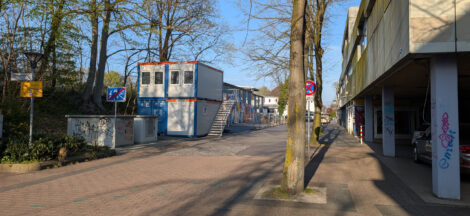 Gestapelte Baucontainer stehen entlang eines Bahndammes auf Parkplätzen gegenüber einem heruntergekommenen Einkaufszentrum.