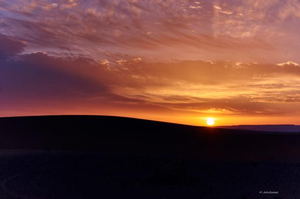 A sky full of hues of orange and yellow punctuated by pockets of blue.  The dunes are dark, silent silhouettes with a background of vibrant colour.
