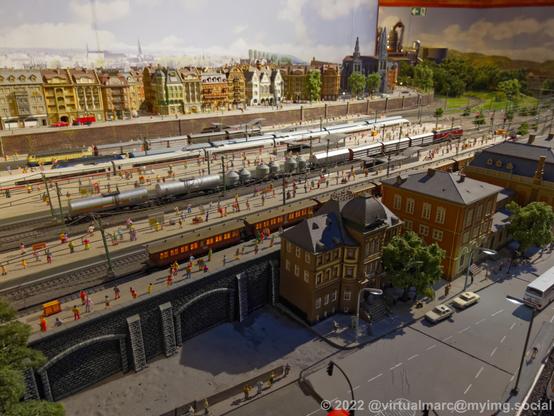 A photo of a big model railway train station with multiple passenger trains of all eras and fright trains driving through it. There are hundreads of passengers on the platforms. There is a city in the back and frot of it, with streets and cars.