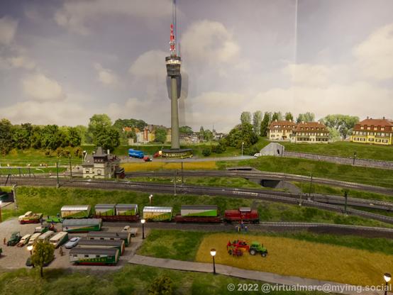 A photo of a model railway with a shunting train on it, transporting wagons of a circus. There is a TV Tower in the back.