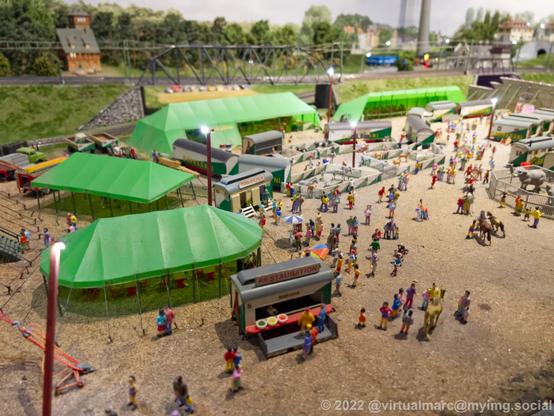 A photo of a circus on a model railway with a restaurant, a toilet and multiple tents and animal enclosures. There are many visitors on this place.