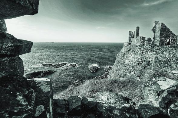 A monochrome photograph shows the ruins of an old stone castle perched on a cliff overlooking the ocean. Waves crash against the rocks below.