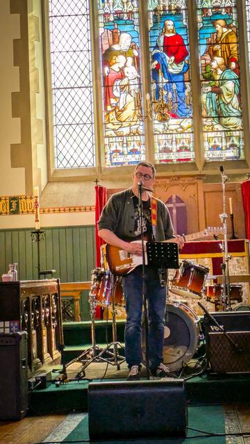 A man on stage with a guitar, behind him is a stained glass window