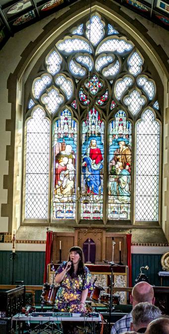 A woman on stage in a glitterly dress, behind her is a stained glass window