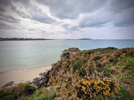 La côte avec l'océan, une plage, des rochers et un chemin herbeux. Le ciel est orageux mais encore beau.