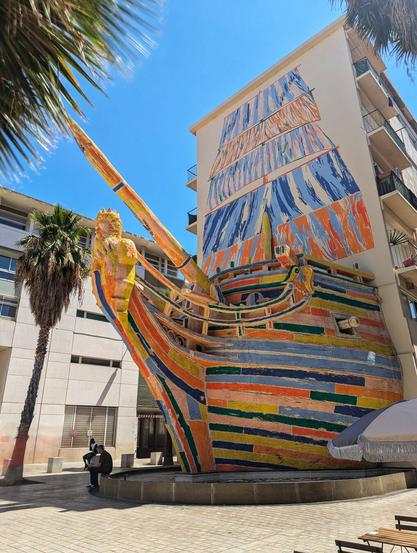 A vibrant and whimsical sculpture of the prow of a ship juts out from the side of a tall, light-coloured building under a bright, clear blue sky. The ship's hull is painted in bold, horizontal stripes of various colours including orange, blue, yellow, and green, giving it a playful, almost Lego-like appearance. The figurehead, a golden-yellow sculpted woman, is prominently displayed at the very front. A long, angled bowsprit extends outwards, painted in shades of yellow and blue.
Above the sculpture, the building's facade features a painted mural depicting the sails of the same ship, rendered in swirling patterns of blue and white, creating a sense of movement. Palm trees frame the left side of the image, adding a touch of tropical ambiance. Below the sculpture, a paved area with benches and a partially visible parasol suggests a public space where people can sit and admire the artwork. A few individuals can be seen in the background, giving a sense of scale to the large installation.