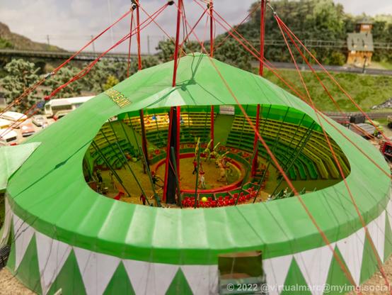 A photo of a circus tent on a model railway. The roof is open so you can look inside, there is currently a show going on.