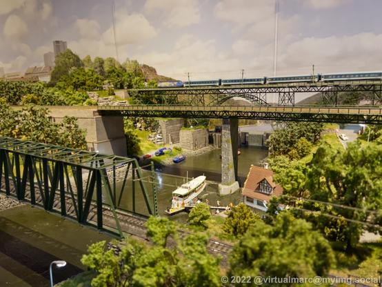 A photo of a river on a model railway. There is a ship for passengers on the river, docket to load/unload passengers. There are three railway bridges going over the river. On the most distant one is currently driving a passenger train.