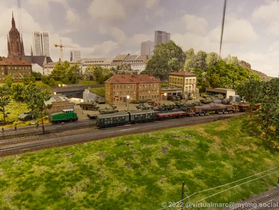 A photo of a military complex on a model railway, with tanks and military trucks. In front of it are railroads with a freight train currently passing by.