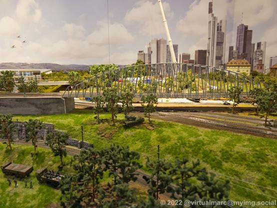 A photo of a model railway with a bridge and som trees and grass. Theres a tank, twi military trucks and some soldiers on it.