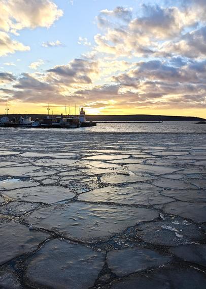 Picture of a lake with broken ice plates on the foreground while sun is setting behind horizon. Sky has light yellow and blue colors with clouds creating some darker colors. On the horizon level there is a slice of land with small lighthouse type building and some boats at the shore.