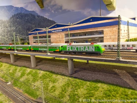 A photo of a model railway. A Volkswagen factory is in the back with a railroad in the front. Two passenger trains are passing by, the front passenger train is green and labled with the FLIXTRAIN Logo.