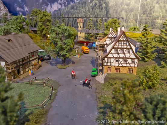 A photo of a small cottage on a model railway with half timbered houses, some old cars, a dump truck and two people riding horses. Theres also a tractor behind a house.