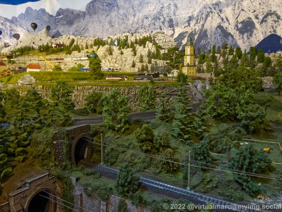A photo of the country side of a model railway with rail tracks, woods, busses and some houses in the back. Theres a hot air balloon flying in the back left.
