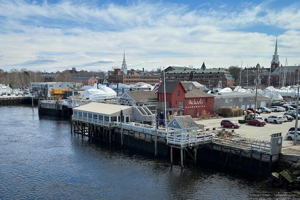 A waterfront scene with a pier extending into the water. Several buildings, including one with a large sign that reads ‘Market,’ are visible. In the background, a town with various buildings and church steeples is seen against a partly cloudy sky