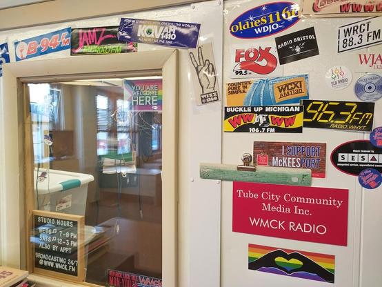 Photo depicts the wall of a radio station decorated with vintage and new bumper stickers reading, "B-94FM," "MIX JAMZ," "KQV 1410," "Oldies 1160," "WCXI" and other names.