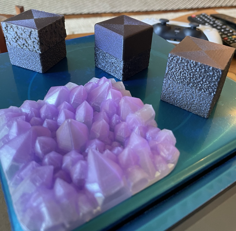 A series of blue/purple 3d printed cubes. In front of the cubes is a 3d print of simulated crystals printed in clear and purple.