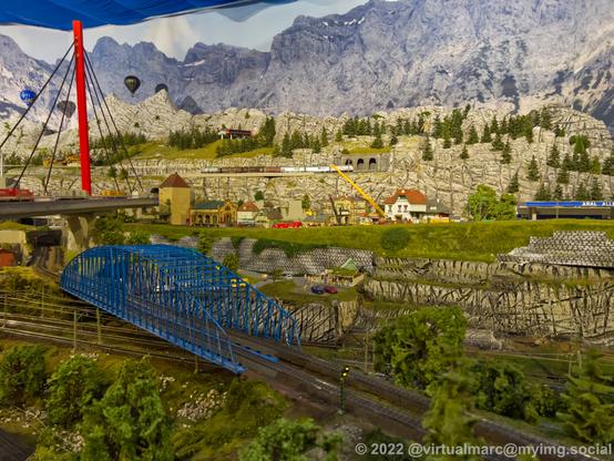 A photo of a model railway with crossing rails over a blue bridge. A red bridge for cars crosses all the rails with trucks on tit. Theres a small village under construction in the back. Two hot air balloons are flying in the air and a cablecar can be seen in the back.