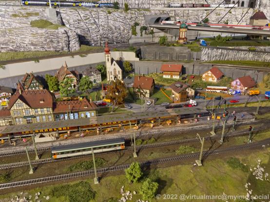 A photo of a small train station of a small village in a model railway, with a passenger wagon standing on a rail. There are cars, trucks and a bus on the roads in the village full of half-timbered houses. Theres also a church.