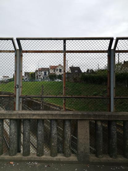 A concrete bridge with a protective fence over train tracks. The train tracks are sided with an upward slope with houses one the top of it.