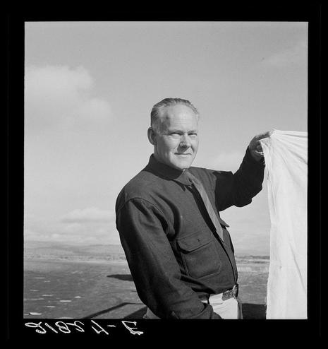 The image depicts a man standing on an elevated surface with the sky and landscape visible in the background. He is dressed in work attire, including a dark jacket, trousers, and boots, suggesting he may be involved in some outdoor or industrial activity. The most striking feature of this black-and-white photograph is that the man is holding up what appears to be a piece of fabric or material with both hands; his expression seems contemplative as he gazes at it.

The text on the bottom right corner reads "81594-F," which could possibly indicate either a date, location code, reference number for an archive collection, or another form of categorization. The overall composition and style of this photograph suggest that it might be part of a larger documentation project related to rural life, work conditions during the mid-20th century in America, particularly connected with agricultural laborers.

This image is likely associated with Dorothea Lange's collection known as "Untitled photo," which was captured by her for the Farm Security Administration (FSA) mobile camp program. The FSA aimed to provide a record of rural life and working conditions during this era through its photographic documentation initiatives, such as documenting Camp Managers or individuals in their work environments.

It is possible that this photograph captures a moment from one of these camps where Lange photographed various aspects of farm laborers'  [...]