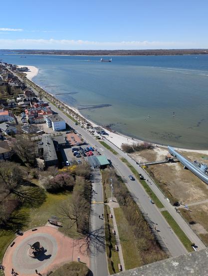 Semi-aerial view of a small seaside town next to a body of water. separated by a road and a beach with white sand. To the far right there is a U-boat on land that is a historic artifact. The weather is sunny, a few low clouds spot the clear spring sky.