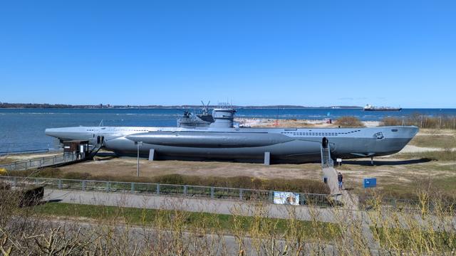 View of a U-boat next to a body of water. There's bushes in the foreground, the submarine dominates the mid distance, the water (which could be a wide river) behind it. On the water a large cargo vessel is on its way to the open sea. The weather is sunny, a few low clouds spot the clear spring sky.