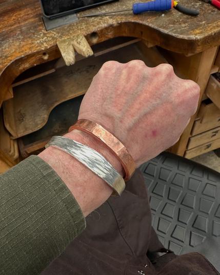 A close-up of a hand wearing two metal bracelets: one copper, and one textured silver. The hand is above a wooden workbench, with tools and materials visible in the background.
