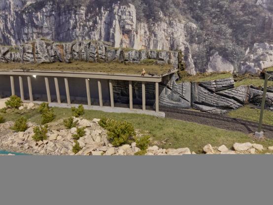 A photo of a half tunnel on a model railway with a small river in the front. There is sitting someone on top of the tunnel.