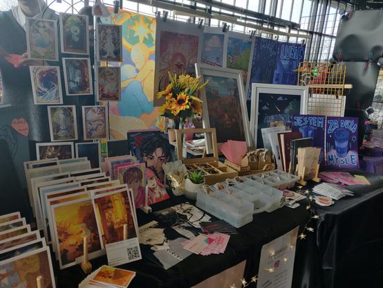 A view of an artist alley table, featuring a variety of artworks in different sizes and style. There is a sunflower bouquet and some fairy lights