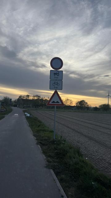 Ein Verkehrsschild mit einem No-Entry-Symbol neben Straßenschildern, die darauf hinweisen, dass Traktoren und Fahrräder erlaubt sind. Der Hintergrund zeigt einen Weg, grasbewachsene Flächen und ein Feld mit einem Sonnenuntergangshimmel.