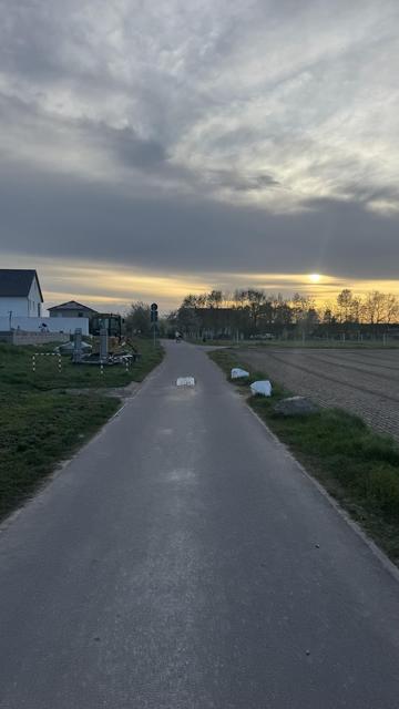 Ein gepflasterter Weg führt zu einem Sonnenuntergang, flankiert von Grasflächen und felsigen Rändern. Auf der linken Seite befinden sich zwei weiße Gebäude, eines mit einem Spielplatz in der Nähe. Im Vordergrund sind Baumaschinen zu sehen. Der Himmel ist bedeckt.
Auf der Straße ist in der Mitte ein Block, der für KFZ die Durchfahrt versperrt.