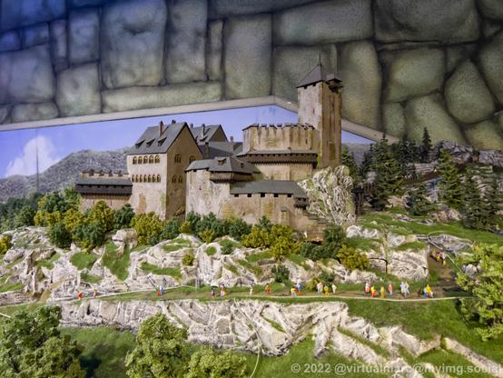 A photo of an old castle on top of a hill on a model railway. A walkway is going up and multiple people are hiking up and down.