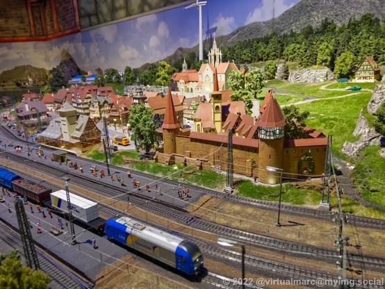 A photo of a model railway with a train station that is passed by a freight train. Behind the train station is a historic city wit ha small castle.