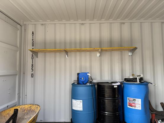 The inside of a shipping container with a shelf mounted on the wall and three 50 gallon drums below it
