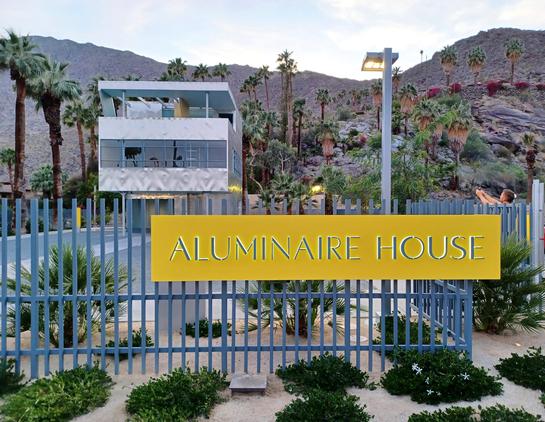A three-story, aluminum-clad, boxy building stands against a background of the brown, rocky San Jacinto mountains and palm trees. In the foreground is a metal fence with a bright yellow sign attached that says "Aluminaire House." The sun is setting behind the mountains at dusk.