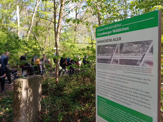 Infotafel "Barackenlager" aus der Reihe "Grüne Infrastruktur - Gremberger Wäldchen" mit historischen Erläuterungen und Luftbildern, im Hintergrund Teilnehmer*innen der Gedenkveranstaltung