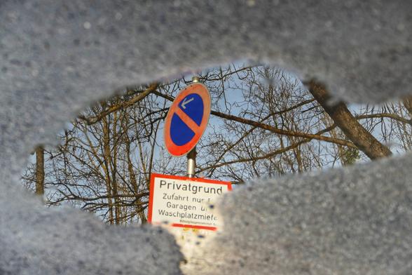Das Bild zeigt ein Verkehrsschild, das sich in einer Pfütze spiegelt. Das Schild ist blau mit einem roten Rand und einem roten diagonalen Strich von oben links nach unten rechts. In der Mitte des Schildes zeigt ein weißer Pfeil nach links. Unter diesem Schild befindet sich ein weiteres Schild mit einem Hinweis, der lautet: „Privatgrund Zufahrt nur für Garagen und Waschplatzmieter Wohnungseigentumsgesellschaft.“ Der Hintergrund des Bildes zeigt kahle Bäume, was auf eine winterliche Szene hindeutet. Das Bild wurde um 180 Grad gedreht und vertikal gespiegelt, sodass es so wirkt, als könnte man wie durch ein Tor (das Äußere der Pfütze) zu diesem Schild gelangen.