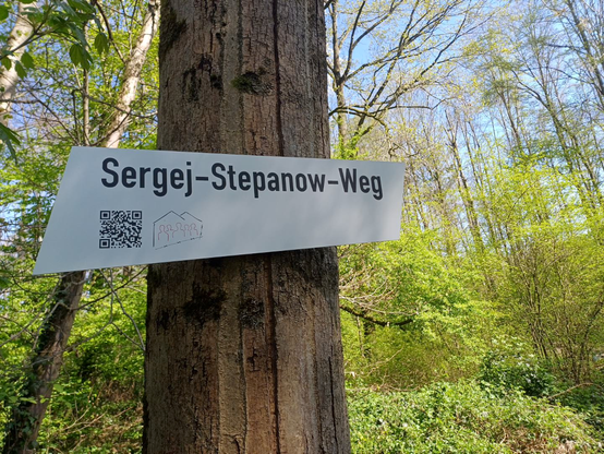 Schild "Sergej-Stepanow-Weg" mit QR-Code zum Audio-Projekt, im Hintergrund hellgrüne Laubbäume im Gremberger Wäldchen