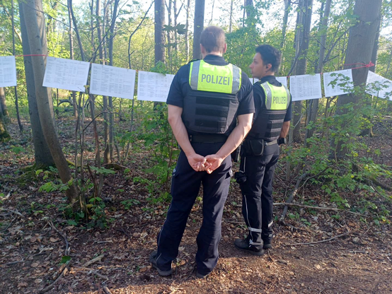 Zwei Polizisten* betrachten die an einer Leine aufgereihten Namenslisten mit den (bekannten) Opfern des NS-Krankensammellagers im Gremberger Wäldchen, im Hintergrund hellgrüne Laubbäume