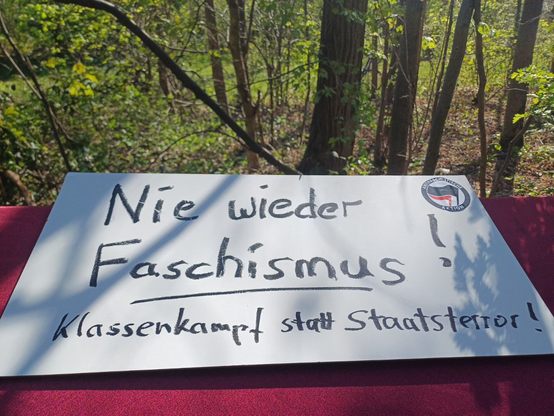 Pappschild: "Nie wieder Faschismus! Klassenkampf statt Staatsterror"!, mit Aufkleber "Antifaschistische Aktion" (mit schwarzer und roter Fahne), im Hintergrund hellgrüne Laubbäume im Gremberger Wäldchen