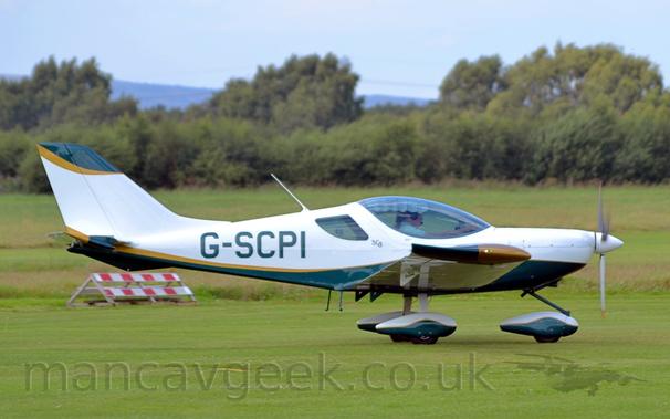 Side view of a white, single engined light aircraft taxiing from left to right on a grass airfield.
The plane is white on top, with a green belly, with a gold stripe separating the 2 colours.
The same colour scheme is repeated on the spats covering the wheels on the fixed undercarriage.
The ends of the wings are gold at the front and green ant the back.
Green grass fills most of the frame, leading up to green grass in the distance, under grey skies.