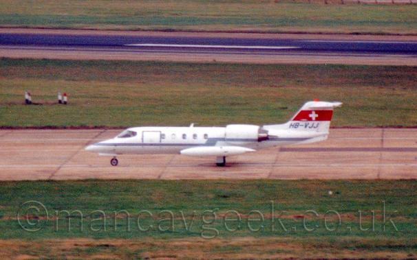 Somewhat grainy side view of a white, twin engined BizJet taxiing along a taxiway from right to left.
The plane is almost entirely white, with a double, thin blue stripe running along the body, and a thick red strip containing a white cross on the tail.
The registration "HB-0VJJ" is under this, in blue.
Over-sized wingtip fuel tanks cover a large part of the lower body.
Green grass fills the bottom of the frame, with more grass and a black runway in the background.