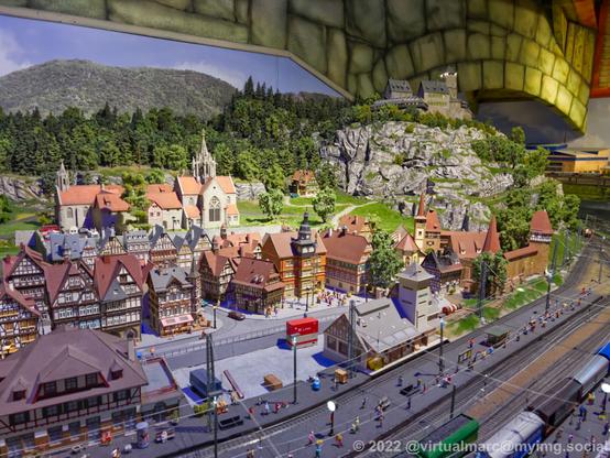 A photo of an old medieval village with half-timbered houses of a model railway. A railway station can be seen at the front, with a freight train. A big church is behind the village and a Castle can be seen at the top of a hill/mountain in the back.