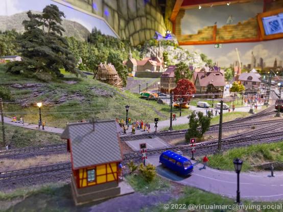 A photo of a railway crossing with people waiting to cross and a car that just crossed of a model railway.