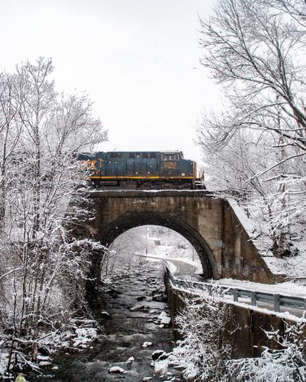 A train crosses a small kestone arch bridge, surround by snow covered trees during a snow storm