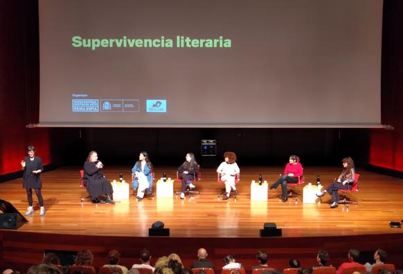 Una imagen de un escenario en un auditorio con una pantalla grande que muestra el texto "Supervivencia literaria". Siete mujeres están sentadas en sillas individuales en el escenario, participando en un panel: Alana Portero, Sara Torres, Ana Flecha Marco, Claudia Muñiz, Bibiana Collado y Sabina Urraca. Una mujer adicional está de pie frente al panel, interpretando en lengua de signos.